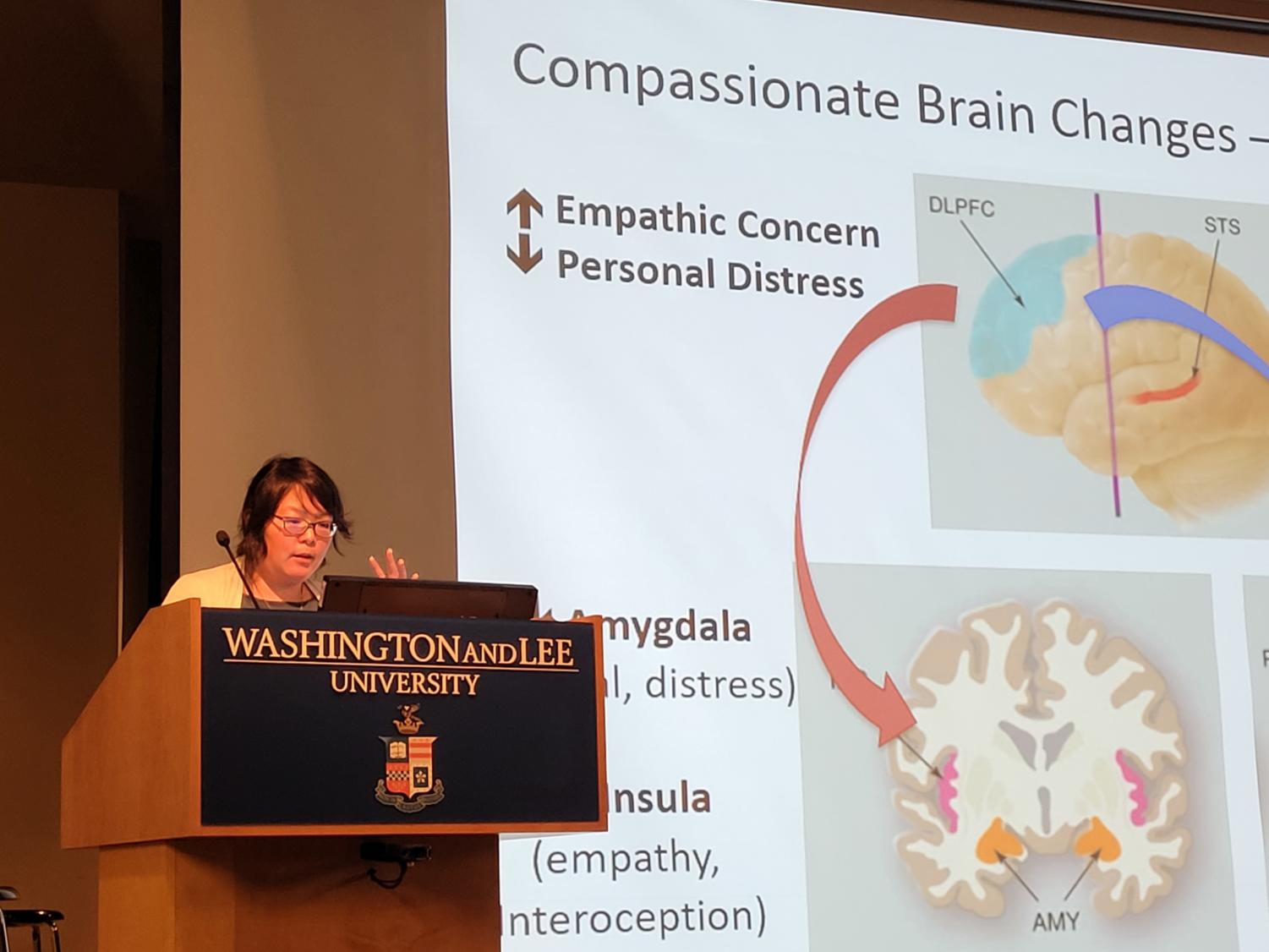 Dr. Helen Weng stands at a podium, gesturing and speaking. Behind her is a screen with the words "Compassion Brain Changes" with diagrams of a brain titled "Empathetic Concern" and "Personal Distress."