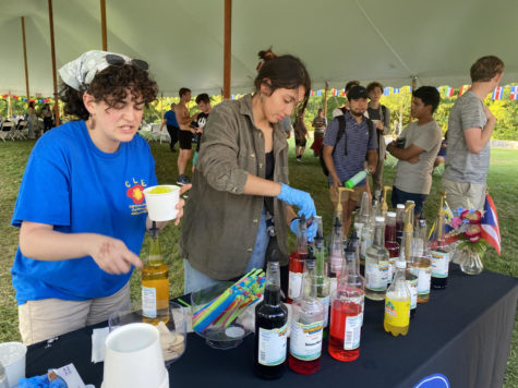 Two students pour brightly colored snow cone flavoring into plastic cups. Students form a line next to the table.