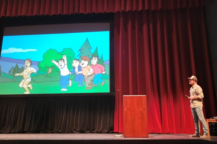 Mike Henry stands on stage in front of a red curtain, looking up at a screen displaying Family Guy images.