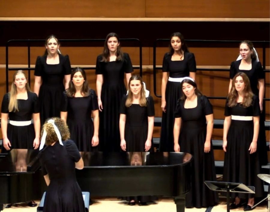 Two rows of women singers dressed in matching black dresses stand behind a piano. In front of them is another student in a black dress, with her back turned towards the camera, as she conducts the singers.