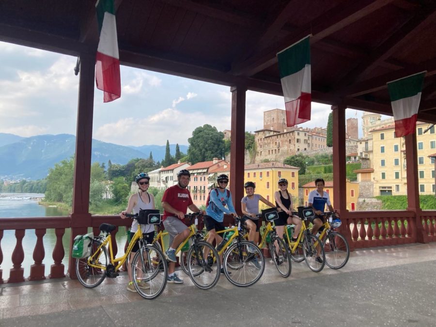 A row of six bikers sit parked on a bridge, with Italian flags lining the roof. In the background, there are multiple colorful buildings and a body of water.