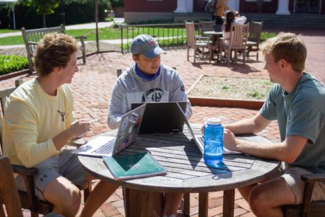 Three students sit around a wooden table outside, two of them holding laptops. They are laughing and talking.