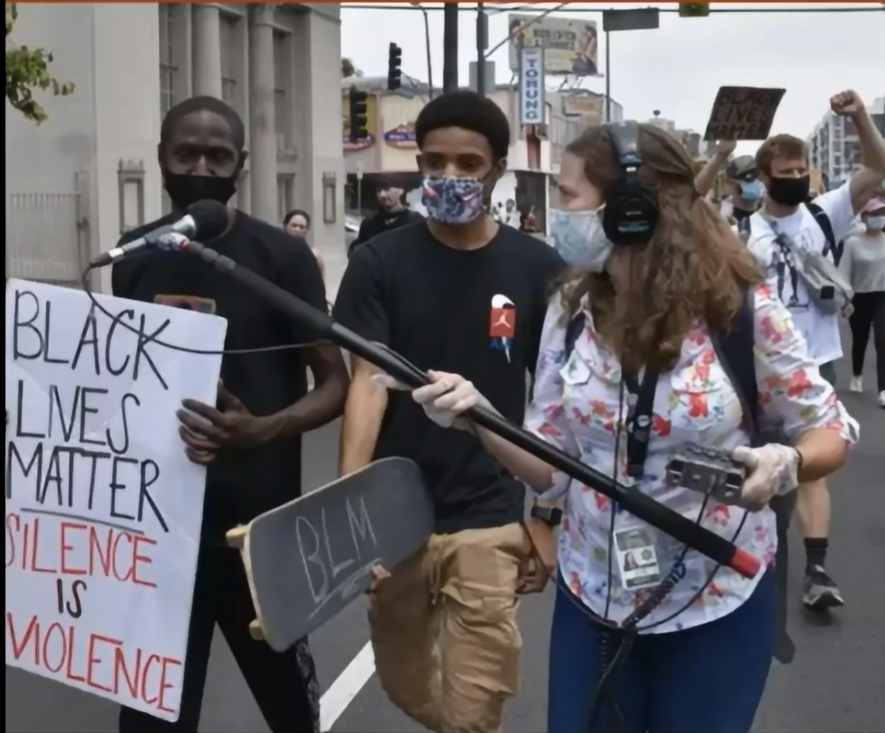 Reporter in a mask and floral shirt holds a boom microphone out to two men holding Black Lives Matter signs in a protest march.