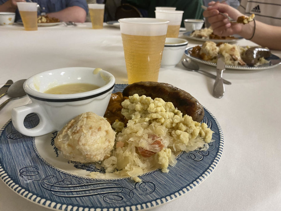 A blue-rimmed plate holds a biscuit, sausage, and other Austrian food items. There is also a teacup on the plate. In the background is a clear plastic cup with light beer.