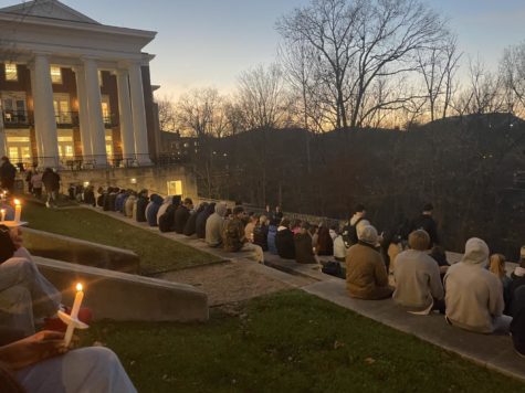 Students sit on concrete stairs with grass in between, holding candles. In the background, there is a lit W&L building.
