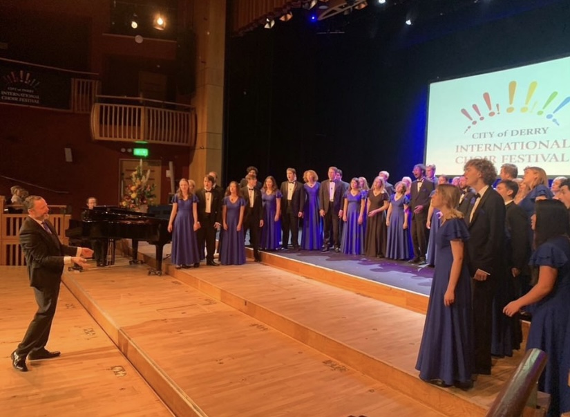 Choir stands on stage in semi-circle wearing blue dresses and black suits. Director stands in front, waving his arms.