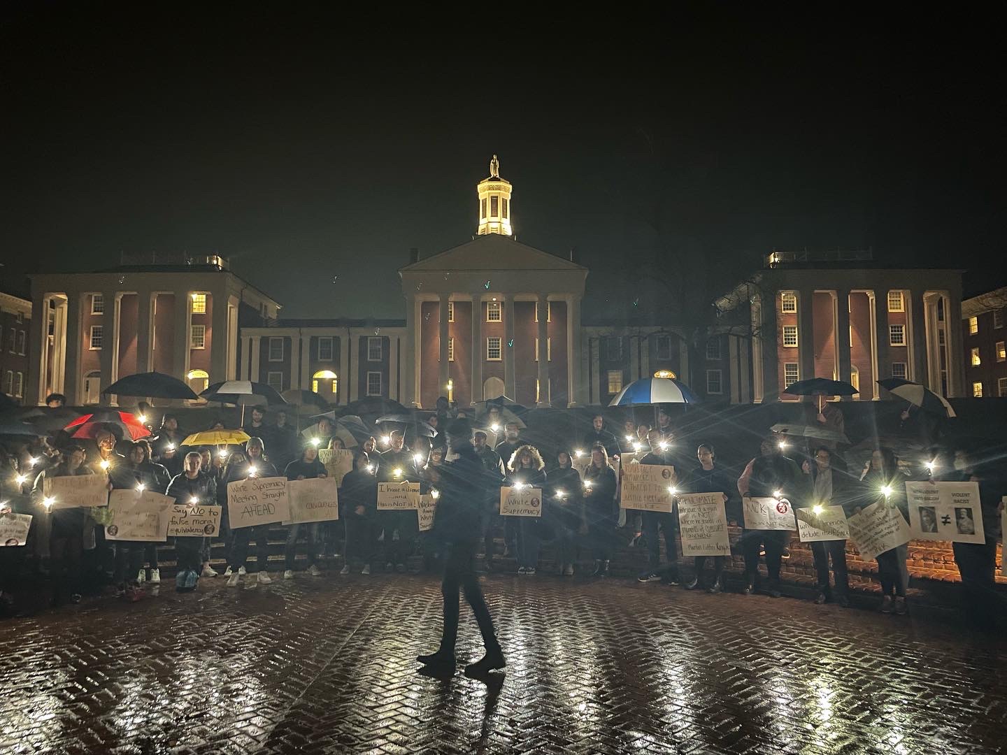 A crowd of people stand in a circle holding flashlights and signs. Behind them are lit up buildings with tall columns.