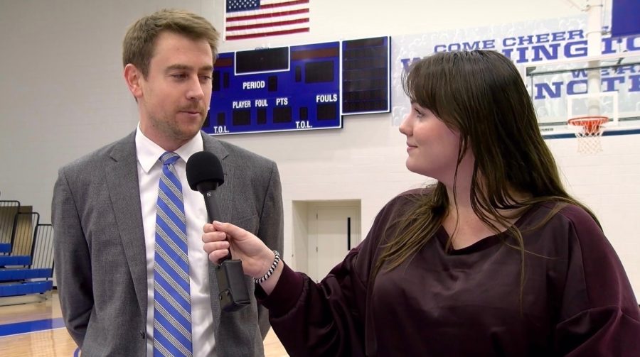 A+woman+holds+a+microphone+in+front+of+a+man+who+is+wearing+a+suit.+They+are+in+a+gym+with+a+basketball+scoreboard.