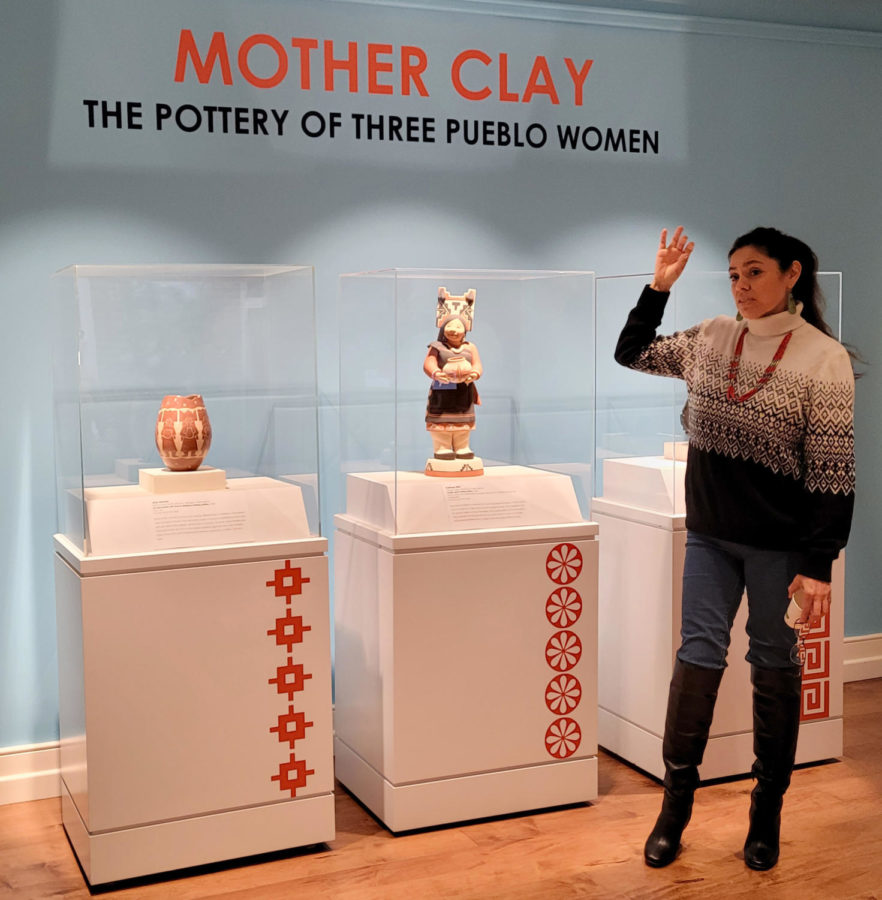 A woman gestures towards a pottery figure in a display case. Two other pottery artworks sit to either side.