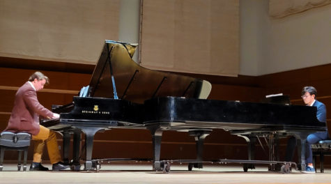Two pianists sit at two pianos, facing each other, on a big stage.