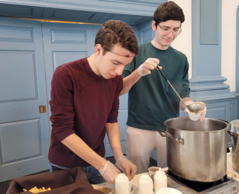 Two young men wearing hair nets ladle soup from a pot into small cups.
