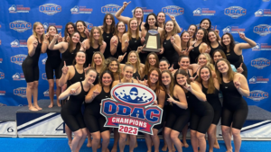 A group of women in black swimsuits smile at the camera. A few women in the front row are holding an ODAC logo.