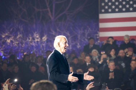 A man with white hair in a suit coat stands above a crowd, speaking. The American flag is in the background.