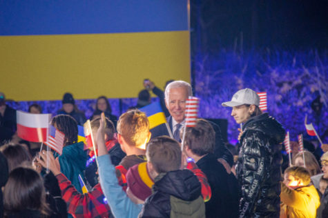 A white haired man in a suit smiles towards a group of children waving American and Ukrainian flags. A large Ukrainian flag is behind him.