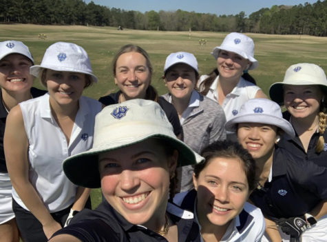 A group of women in white golf outfits and hats smile at the camera. They are on a golf course.