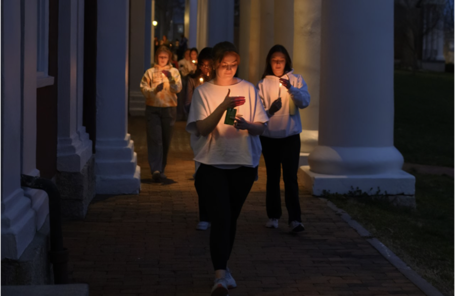 A group of people, appearing mostly female, hold lit candles and walk in a loose line on a brick pathway under columned porches. The photos lighting is dim and indicates twilight.