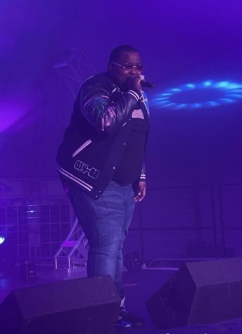 A Black man wearing a letter jacket and jeans sings into a microphone on a stage. The stage lighting is blue.