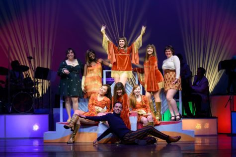 A group of young women, all wearing different 1960s outfits, pose onstage and smile. One young man wearing black sits on the ground and smiles as well.