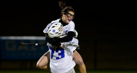 A woman grinning and wearing sports goggles jumps into the arms of another woman in a white goalie helmet. Both are in white and blue uniforms.