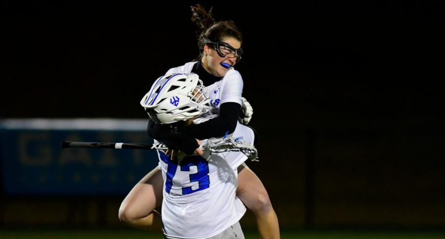A+woman+grinning+and+wearing+sports+goggles+jumps+into+the+arms+of+another+woman+in+a+white+goalie+helmet.+Both+are+in+white+and+blue+uniforms.