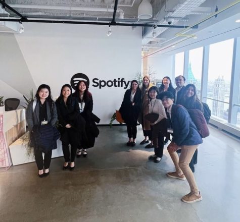 A group of people in business casual outfits pose in front of a wall sign that says Spotify, in a building with a concrete floor.