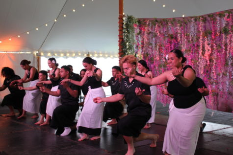 A group of male and female dancers in black and white Polynesian outfits half-kneel and spread their arms toward the audience.