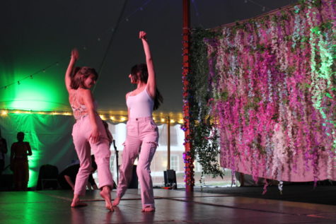 Two women in white tops and khaki pants dance facing each other on a stage. They each raise their right hand and twist their hips and legs. The background has a hanging screen with decorative flowers.