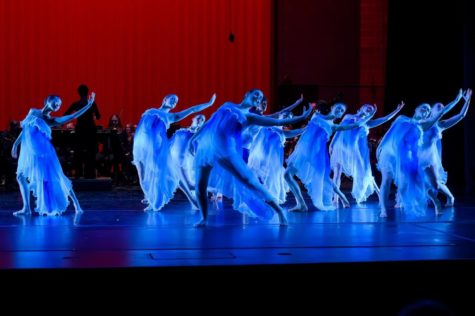 Dancers in draping costumes bend backward and stretch out arms in unison under blue stage lighting.