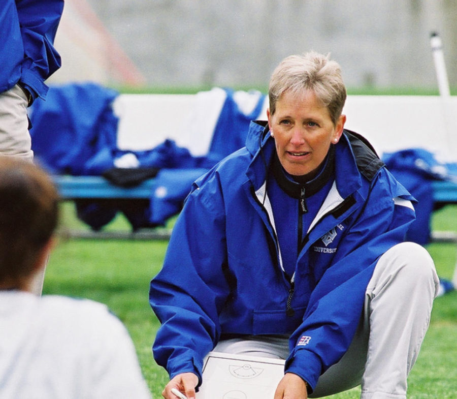 A woman in a blue windbreaker kneels on the sidelines of a sports field. She holds a clipboard and speaks to a group of seated players.