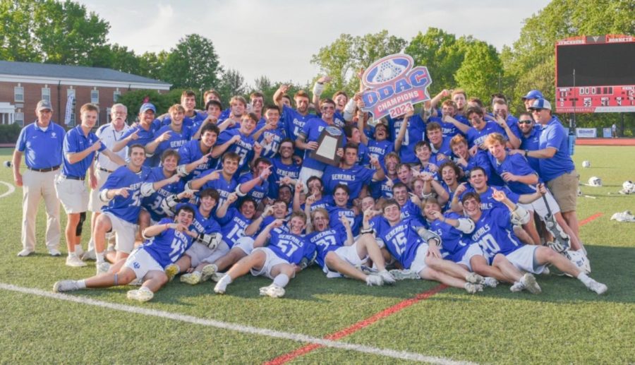 A+large+group+of+lacrosse+players+in+blue+uniform+tops+and+white+shorts+pose%2C+along+with+coaching+staff+in+similar+colors.